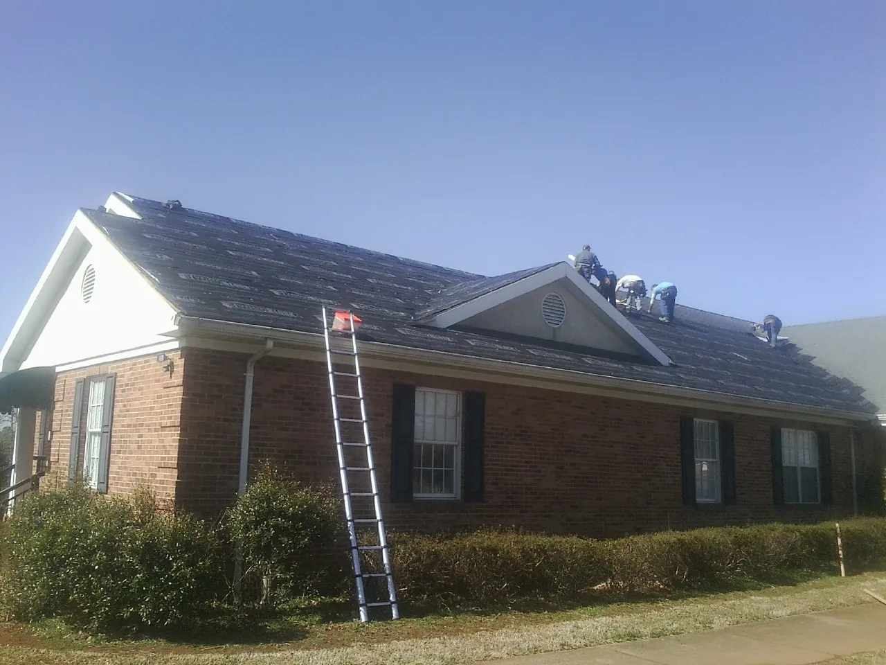 Crew tearing shingles off a brick ranch roof with a ladder set — Metal Roof Repair work by our crew in Hazelton