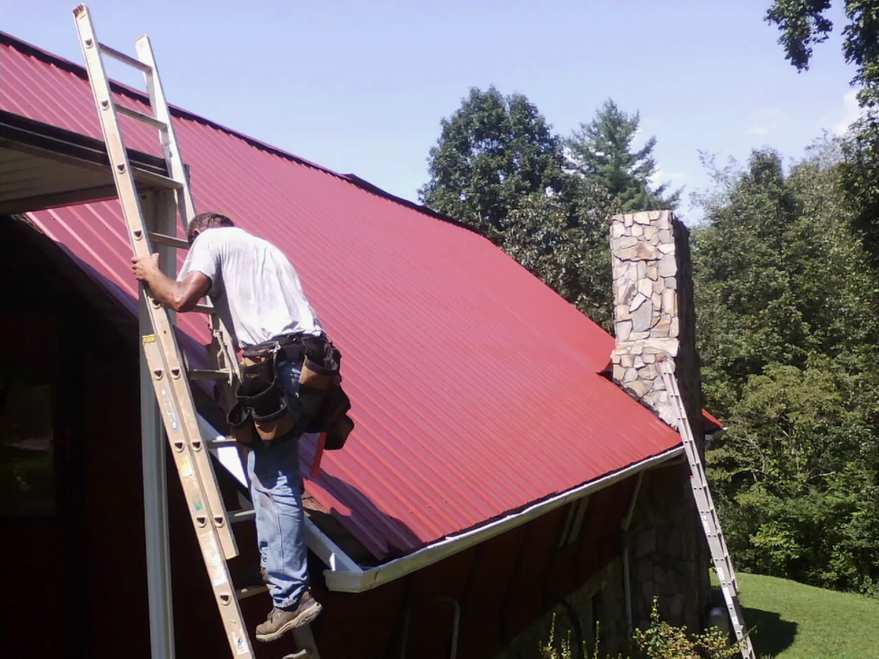 Roofer on a ladder beside a red metal roof and stone chimney — Soffit & Fascia Repair by Roofing Brothers in Hazelton